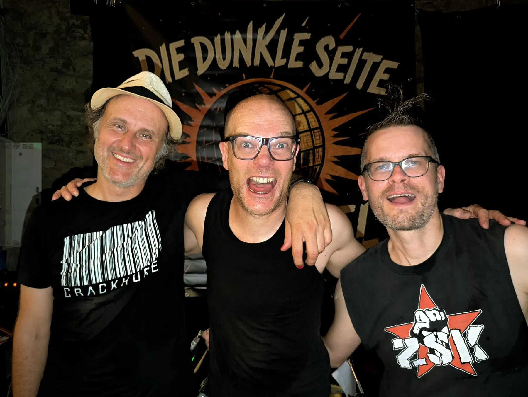 Die Bandmitglieder von 'Die dunkle Seite der Discokugel' vor ihrem Band-Banner: Auge (links, mit hellem Hut), Bebo Softmachine (Mitte, ruft energiegeladen in die Kamera) und Tobir Trichter (rechts, mit auffälliger Punk-Frisur)