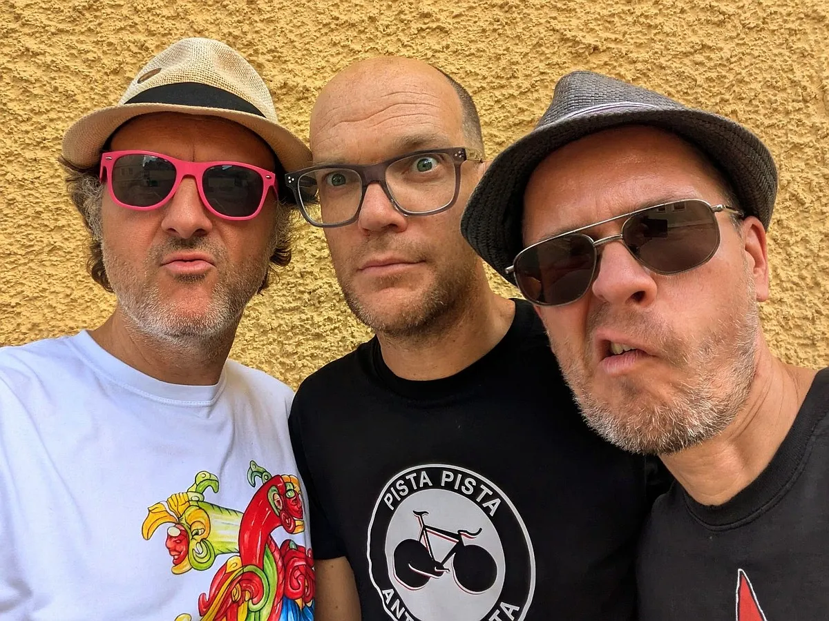 Die Mitglieder der Punk-Band 'Die dunkle Seite der Discokugel' vor einer gelben Wand: Auge (links, mit pinker Sonnenbrille und Strohhut), Bebo Softmachine (Mitte, blickt mit großen Augen direkt in die Kamera) und Tobir Trichter (rechts, mit grauem Hu